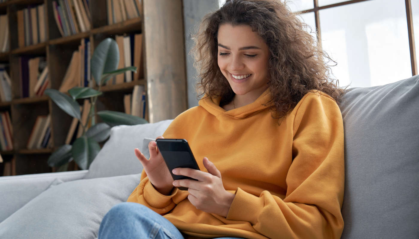 Eine lächelnde junge Frau sitzt gemütlich auf der Couch und nutzt ihr Smartphone.