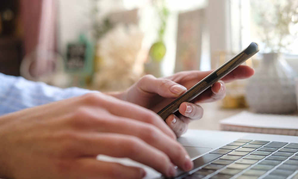 Eine Person sitzt an einem Laptop und hält gleichzeitig ein Smartphone in der Hand. Im Hintergrund ist ein helles, verschwommenes Fenster mit Pflanzen zu sehen.