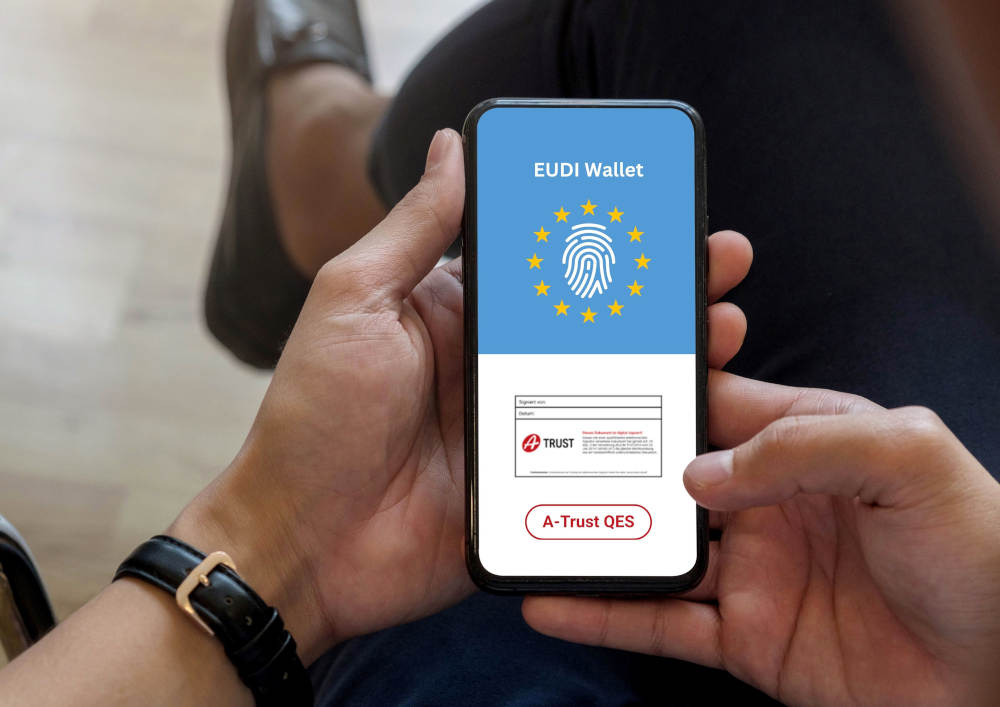 Eine Person hält ein Smartphone; Auf dem oberen Bildschirmbereich steht „EUDI Wallet“ neben einem Fingerabdruck-Symbol in den Farben der Europaflagge; Im unteren Bereich ist die A-Trust Signaturmarke zu sehen sowie ein Button mit der Beschriftung „A-Trust QES“.