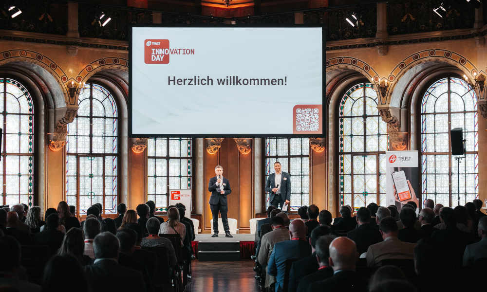 Szenerie: Markus Vesely und Sven Glöckner eröffnen auf der Bühne des Palais Ferstel den A-Trust Innovation Day; Vor ihnen befinden sich vom Publikum gefüllte Sitzreihen.