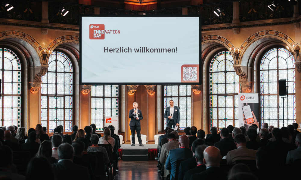 Szenerie: Markus Vesely und Sven Glöckner eröffnen auf der Bühne des Palais Ferstel den A-Trust Innovation Day; Vor ihnen befinden sich vom Publikum gefüllte Sitzreihen.