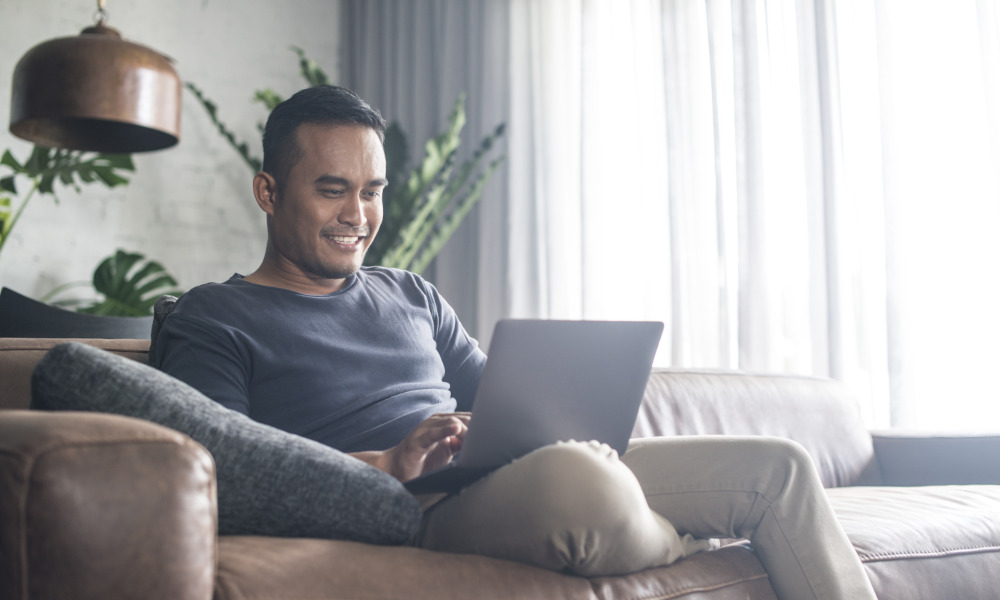 Ein lächelnder junger Mann sitzt entspannt am Sofa und blickt in seinen Laptop.