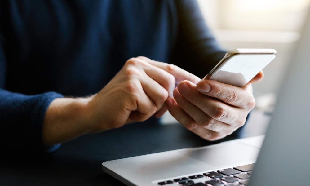 Person mit Mobiltelefon vor Laptop sitzend