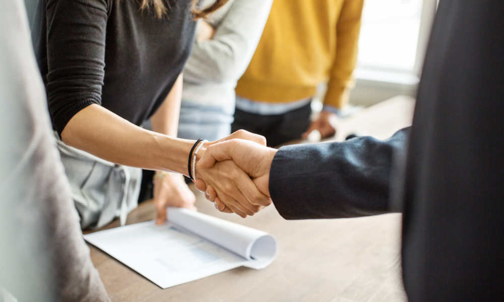 Auf dem Bild sind zwei Personen, die sich die Hände schütteln. Ihre Gesichter sind nicht zu sehen, was den Fokus auf die Geste des Handschlags legt. Im Hintergrund befinden sich undeutlich 2 weitere Personen. Der Handschlag findet über einem Tisch statt, auf dem Dokumente liegen, was darauf hindeutet, dass eventuell eine Vereinbarung oder ein Vertrag besiegelt wird.