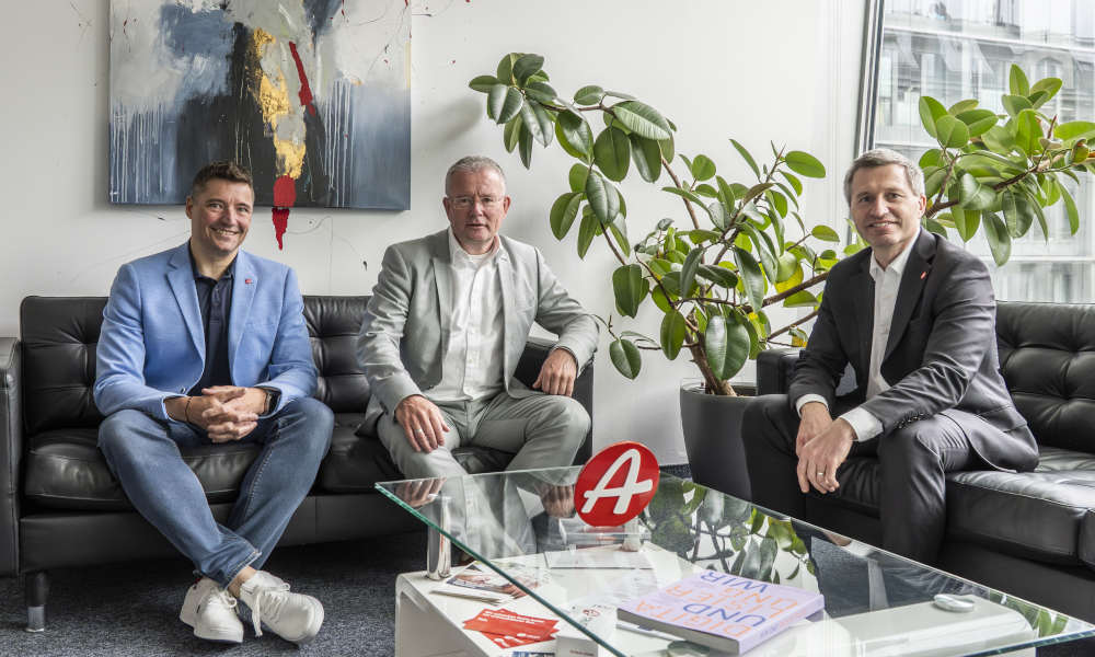 Drei Männer in Anzügen sitzen auf schwarzen Ledersofas in einem modernen Büro mit Glasfront und großen Pflanzen. Auf dem Glastisch vor ihnen liegt ein Buch und ein rotes Logo in Form eines „A“. Im Hintergrund hängt ein abstraktes Gemälde.