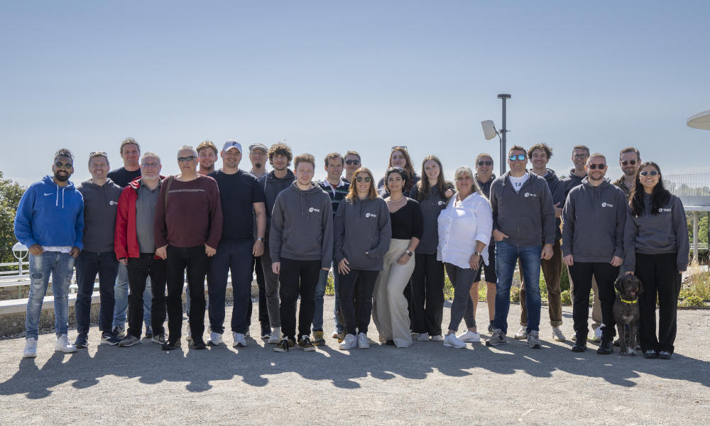 Gruppenfoto der A-Trust Mitarbeiter:innen im Freien bei sonnigem Wetter. Das Team steht nebeneinander und lächelt in die Kamera, einige tragen A-Trust Hoodies. Ein Hund ist ebenfalls Teil der Gruppe.