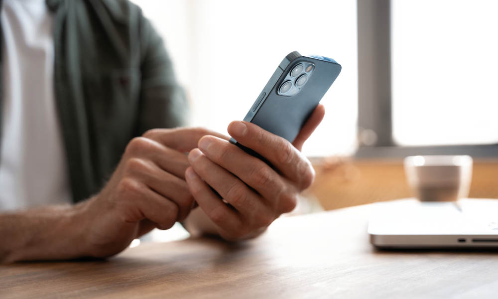 Eine Person sitzt an einem Holztisch und hält ein Smartphone mit beiden Händen. Im Hintergrund ist ein Laptop sowie ein Glas Wasser zu sehen.