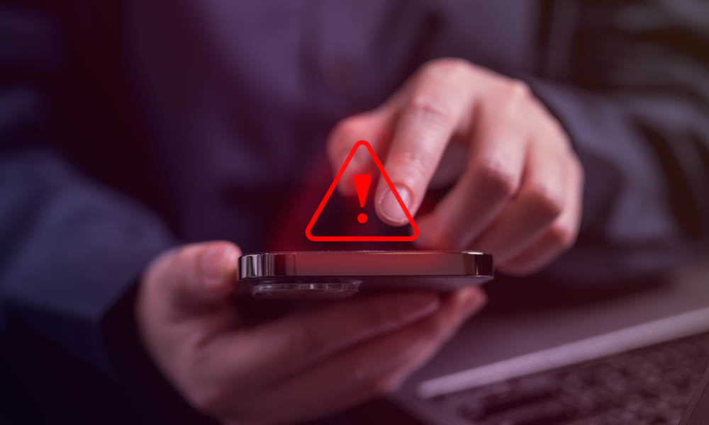 Montage: Eine Person hält ein Smartphone in der Hand und tippt darauf, während ein rotes Warnsymbol mit Ausrufezeichen über dem Gerät eingeblendet ist. Im Hintergrund ist ein Laptop unscharf zu erkennen.