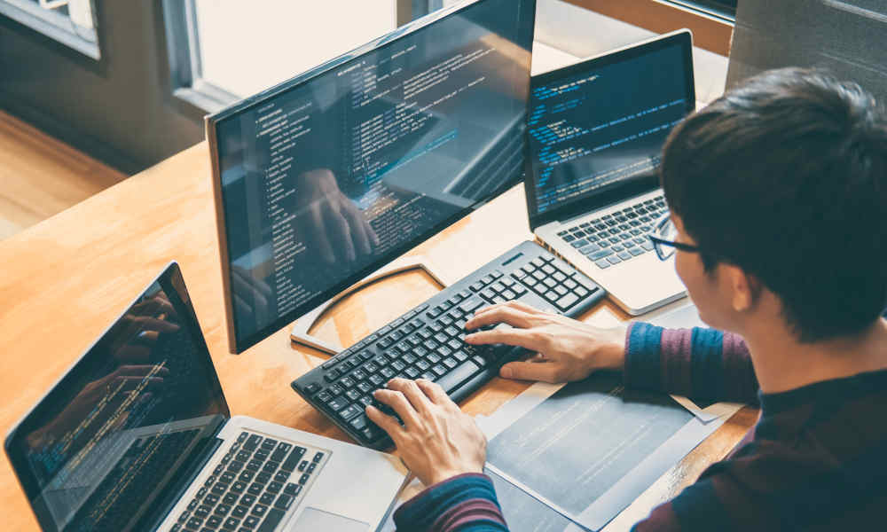 Stimmungsbild: Junger Mann sitzt am Schreibtisch vor zwei Laptops und einem Bildschirm und programmiert.