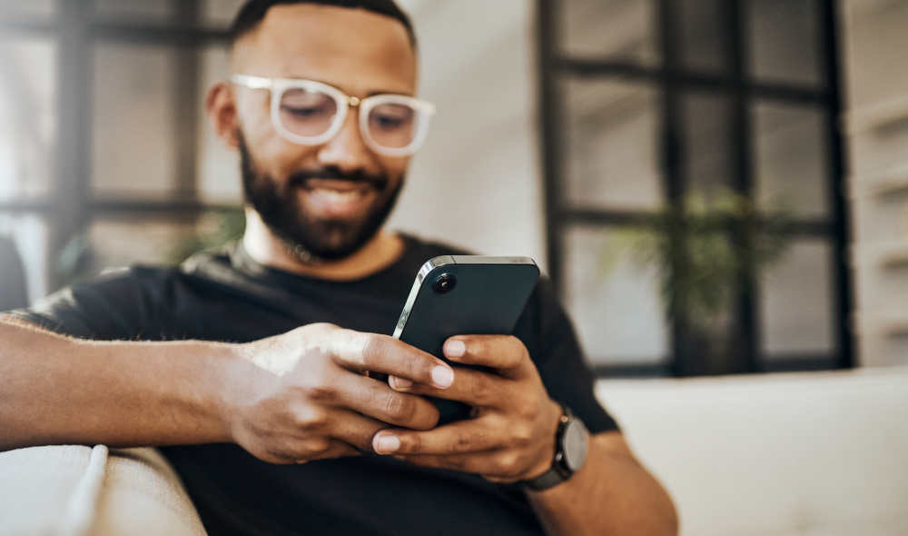 Ein junger, lächelnder Mann sitzt auf der Couch und nutzt sein Smartphone