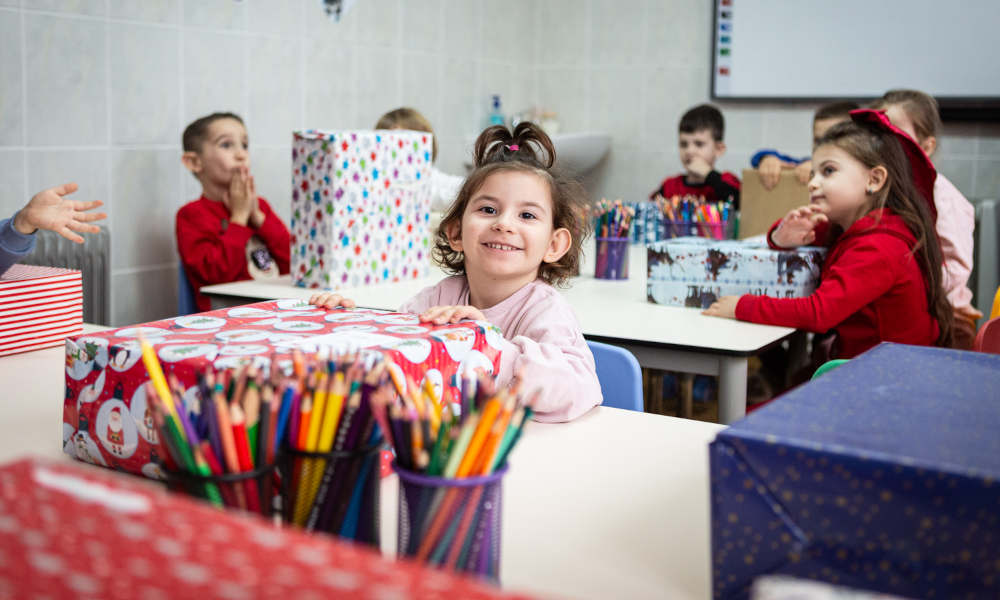 Klassenzimmer mit mehreren Kindern, die Weihnachtspäckchen erhalten haben; Im Vordergrund ist ein kleines Mädchen, das strahlend ihr Geschenk in den Händen hält.